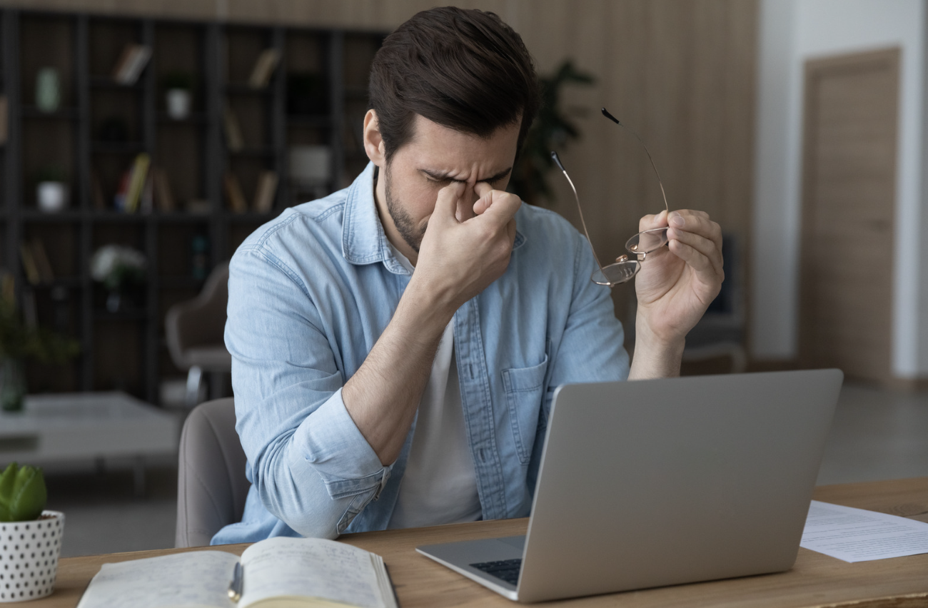 A man taking off his glasses and rubbing the bridge of his nose due to severe eye strain and fatigue while working at a laptop.