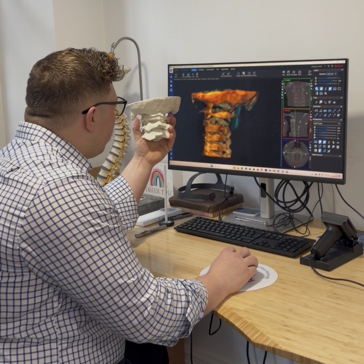A chiropractor analysing 3D spinal imaging on a monitor while holding an upper cervical bone model for precise structural analysis.